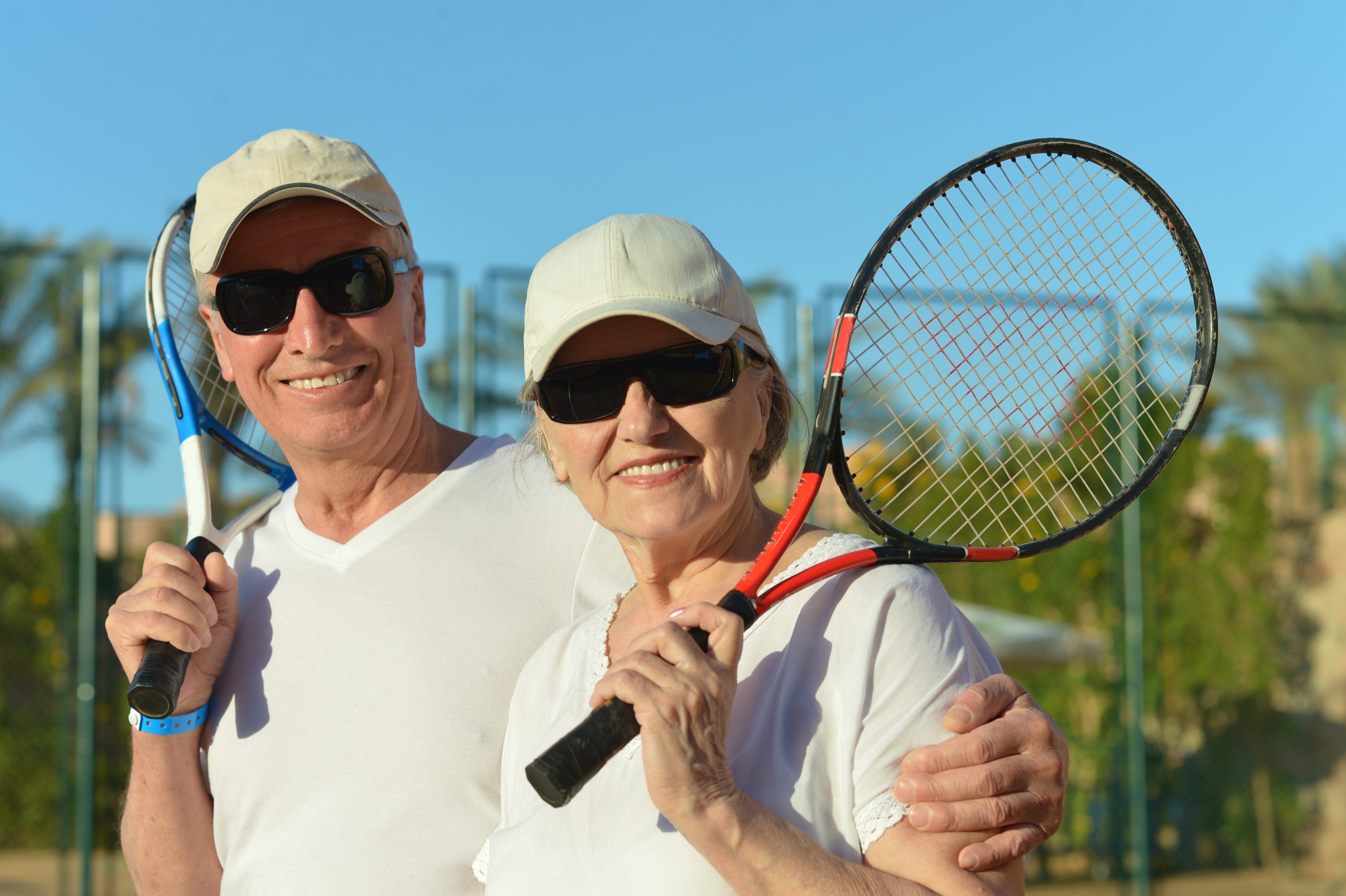 A smiling, happy couple wearing sunglasses rests between sets with tennis rackets in hand.