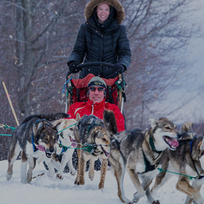 Woman driving dogsled and man sitting in sled pulled by a pack of sled dogs at Treetops Resort.