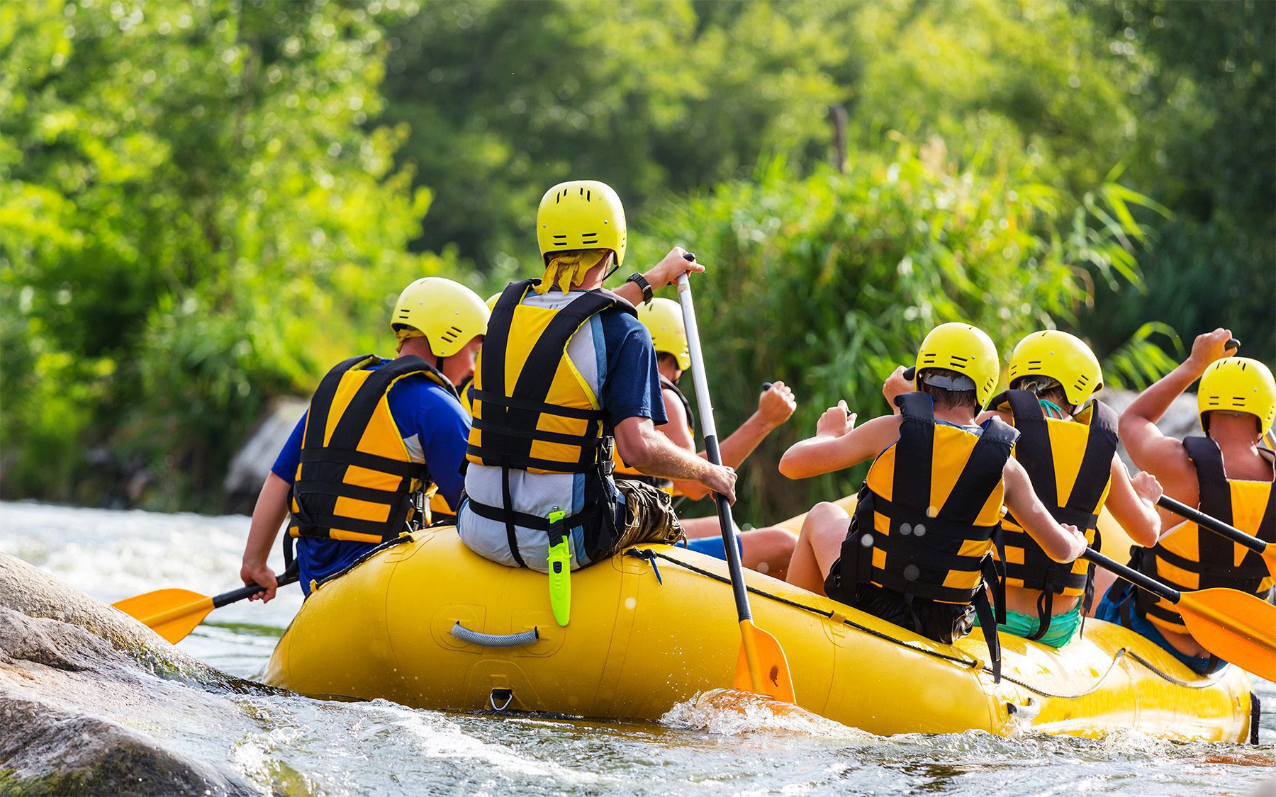 A group of people in full safety gear participate in a rafting excursion on a river.
