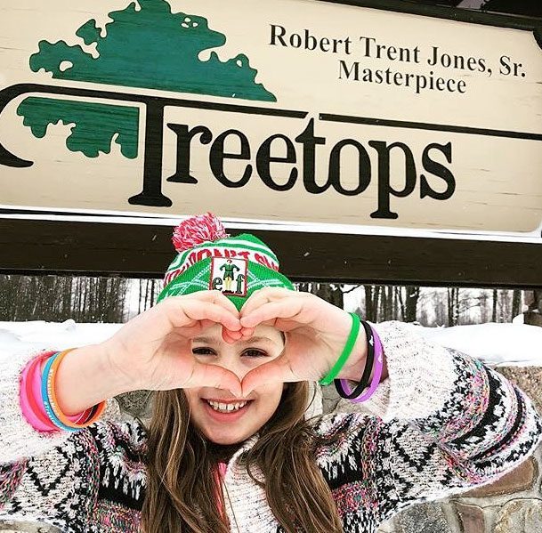 A girl stands in front of a Treetops Resort sign on a winter day, making a heart shape with her hands.