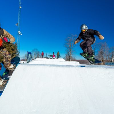 Two snowboarders hit the jumps on a sunny day at Treetops Resort.
