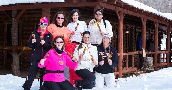 7 women in bright colored winter clothing standing and kneeling together in front of a cabin and holding kabobs 