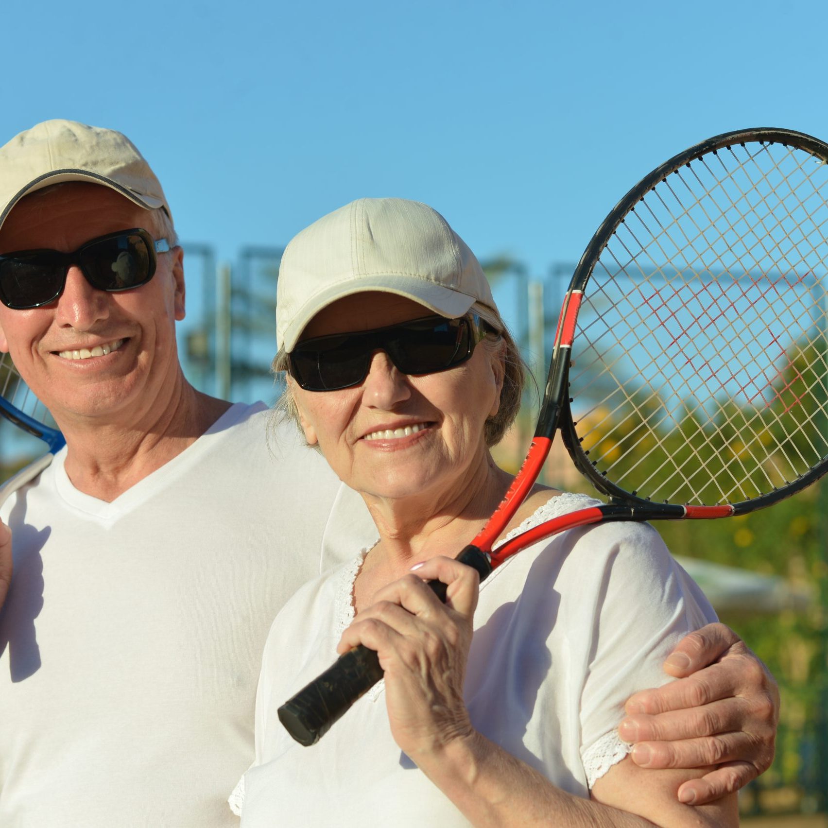A smiling, happy couple wearing sunglasses rests between sets with tennis rackets in hand.